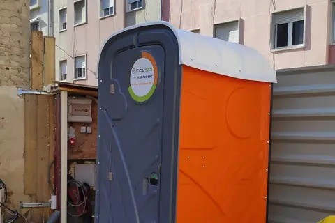 Cabine sanitária INOVSAN laranja e cinzenta instalada numa obra junto a um prédio